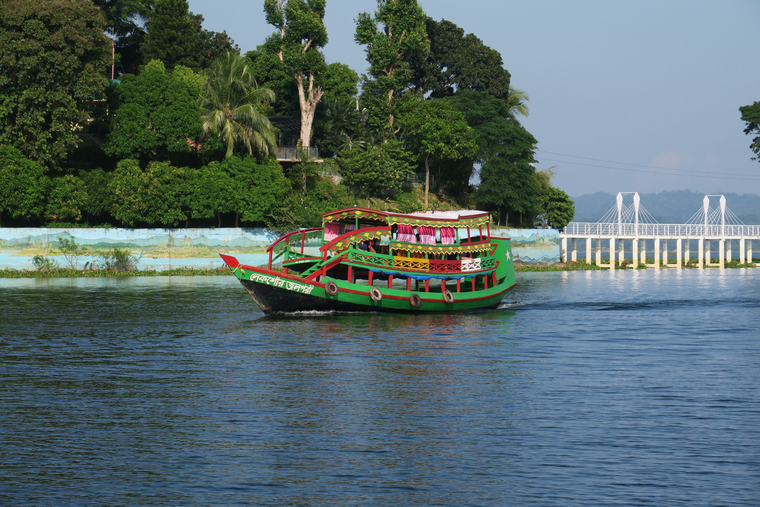 A vibrant green and red boat sails on a calm lake with lush trees and a white bridge in the background.