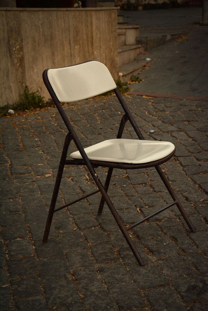 Home A solitary folding chair sits on an empty cobblestone path outdoors.