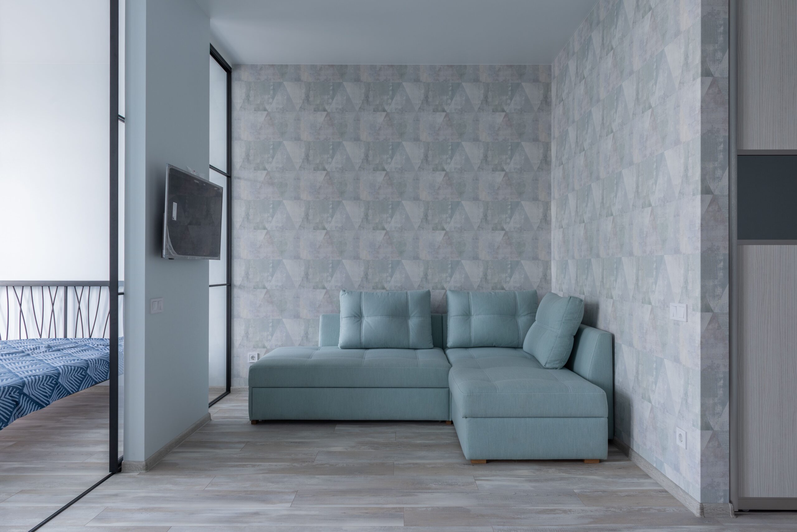 Home Comfortable sofa with cushions placed near wall with wallpaper and bedroom with glass wall and bed in modern flat with TV