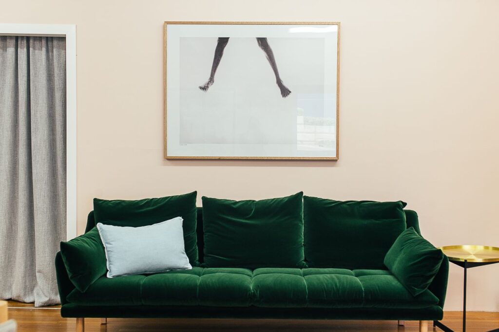 Comfortable green velour sofa with soft cushions and small round table placed on parquet floor near wall with creative framed picture in stylish living room