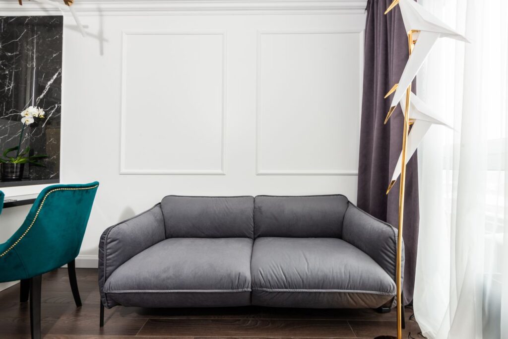 Comfortable gray couch placed at white wall between turquoise chair and window with curtain in contemporary light room with decorations